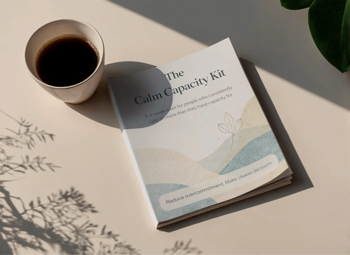 The Calm Capacity Kit booklet shown alongside a cup of coffee, with abstract illustrated hills on the cover and the subtitle 'A 4-week reset for people who consistently take on more than they have capacity for.'